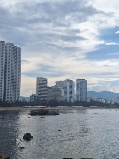 The image depicts a coastal city scene, likely in Nha Trang, Vietnam, characterized by a beach, ocean, and urban development. In the foreground, the ocean occupies a significant portion of the frame, with gentle ripples on its surface reflecting the cloudy sky. Several large rocks are visible, some partially submerged in the water, indicating a rocky coastline or a shallow bay.
The middle ground showcases a sandy beach lined with a dense arrangement of palm trees. Numerous white umbrellas are set up on the beach, suggesting it is a public area for recreation and relaxation. A few small figures of people can be discerned on the beach, engaged in various activities like walking or sunbathing.
Dominating the background is a skyline of modern, multi-story buildings. The most prominent structure on the far left is a very tall, white high-rise apartment building with a uniform facade of many windows. To its right, a series of other hotels and residential buildings of varying architectural styles and heights are visible. Some buildings feature unique designs, such as one with curved balconies. In the distance, beyond the city buildings, a range of mountains can be seen, their peaks softened by the haze and the overcast sky. The sky itself is a mix of light and dark gray clouds, with patches of blue sky peeking through, indicating a possibly humid or transitional weather condition. The overall impression is one of a vibrant coastal city that blends natural beauty with urban infrastructure.