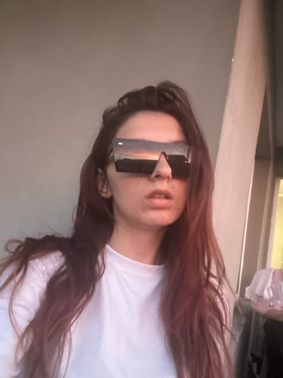 The image is a close-up, waist-up photograph of a young person with long, dark hair wearing large, rectangular sunglasses. The sunglasses reflect a sunset scene with orange and pink hues. The person is wearing a plain white t-shirt. The background is a plain, light-colored wall, with a vertical light-colored beam to the right. A partially visible object with a pink, ruffled texture is in the far right background. The lighting suggests it is either early morning or late evening. The location is not identifiable as being in the city of Tryavna, Bulgaria.