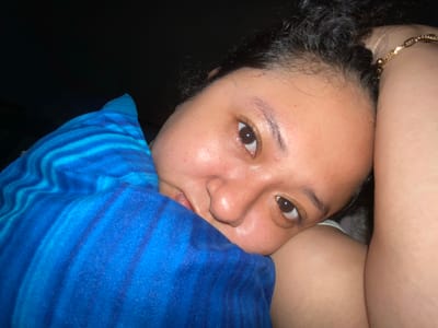 A close-up, overhead shot shows a person lying down with their head resting on their arm, covered by a blue striped blanket. The person's face is partially visible, with one eye looking directly at the camera. Their hair is dark and curly, and they are wearing a gold bracelet on their wrist. The background is dark, suggesting an indoor setting at night. No specific location in Gelam, Indonesia is discernible from the image.