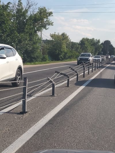 A roadside view of a traffic queue on a clear, sunny day. The image is taken from inside a vehicle looking forward. A white SUV is parked on the left side of the frame, parallel to the road. Ahead, a line of cars is stopped on a two-lane asphalt road. A metal rope barrier with vertical posts separates the lanes. Trees line the right side of the road. The sky is blue with sparse white clouds. The scene appears to be in Chepelyovo, Russia, given the context.