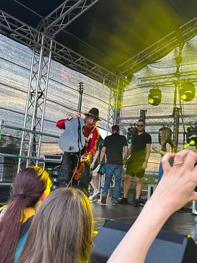 On a brightly lit outdoor stage, a performer in a red shirt and black hat holds a dark gray guitar while a violin is visible below. The stage is constructed from silver metal trusses and draped with white mesh fabric, illuminated by yellow stage lights. Several people are on the stage, including men in black shirts and shorts, and a woman in a black top and jeans. In the foreground, two audience members with their backs to the camera, one with dark red hair in a ponytail and the other with gray hair, are visible. The scene appears to be a live music performance in Tallinn, Estonia, possibly at the Tallinn Song Festival Grounds.