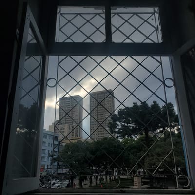 The image shows an open window looking out onto a cityscape with apartment buildings, trees, and a street scene. The window has a diamond-patterned metal security grill. The view includes two large apartment buildings in the distance and closer buildings and trees. There is also a view of a street with cars and pedestrians.

Location: Curitiba, Brazil (based on the style of the buildings and the presence of Araucaria trees, which are characteristic of the region).