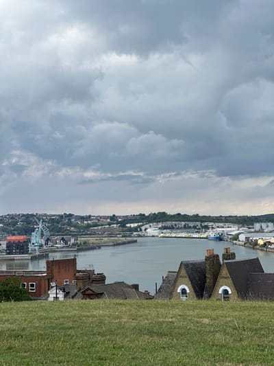 The image displays an elevated view of a section of Chatham, United Kingdom, overlooking the River Medway under an overcast sky.
In the foreground, a field of green grass occupies the lower approximately 25% of the frame. Immediately beyond the grass, partially obscured, are the upper portions of several residential buildings. Two prominent structures in the lower right feature light-colored gable ends, each with a single arched window, and dark, pitched roofs with brick chimneys. A red-brick building is visible further to the left, also partially obscured.
The midground is dominated by the wide, greyish-green expanse of the River Medway, which flows horizontally across the scene. On the left bank, a distinctive large, light blue gantry crane is visible, alongside dark-roofed buildings and what appears to be a red-brick commercial or industrial building. On the right bank, a series of industrial structures, including warehouses, are present. A blue vessel is docked, and long rows of stacked materials, possibly timber or construction supplies, line the quay. Beyond these industrial areas, a significant white chalk cliff or quarry face is visible.
The background consists of a landscape of rolling hills, with scattered residential properties and trees extending into the distance on both sides of the river. The sky is uniformly overcast with shades of grey clouds, indicating diffuse light conditions. Small dark specks, consistent with birds, are observable in the upper left portion of the sky. No individuals are discernible within the scene.