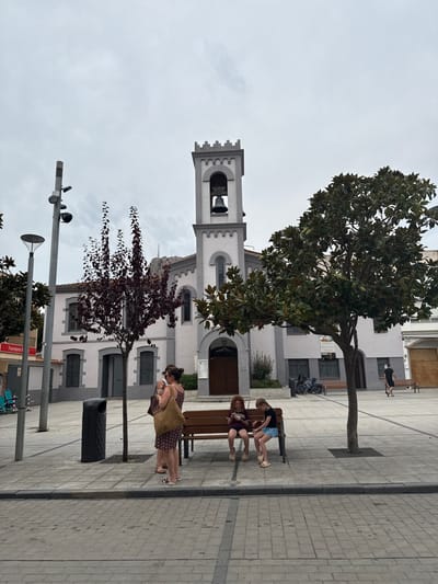 The image depicts an urban plaza in L'estartit, Spain, under an overcast sky. A prominent white and light grey building, featuring a central bell tower with a dark bell and a crenellated top, dominates the background. This structure includes arched windows and a dark wooden entrance with a notice board. The plaza surface is composed of light grey paving stones with darker grey border lines.

In the foreground, two lampposts stand on the left, one equipped with a security camera. A dark grey cylindrical public waste bin is situated near a small tree with dark reddish-purple foliage. To the right, a larger tree with dark green, broad leaves is positioned within the paved area.

Four individuals are visible in the mid-ground. Two adult females stand: one in a patterned dress carrying a woven bag, and a second female slightly behind her. To their right, two children are seated on a dark brown slatted wooden bench, both looking downwards at objects in their hands.

In the background, a person in dark attire walks away on the right side of the plaza. Two bicycles are seen leaning against a building façade to the right of the central building's entrance. An adjacent building to the left of the main structure displays a partial 'Ferreteria' sign.