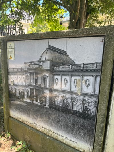 The image shows a weathered outdoor advertisement or information panel displaying a black and white photograph of a grand building. The photograph is framed by a rough concrete border. At the top left of the panel, there is a smaller sign with text that includes "COFFRET ECLAIRAGE PUBLIC" and a serial number "EP0902". There is also a yellow warning triangle with a lightning bolt symbol.

The main photograph depicts an ornate, neoclassical building with a prominent dome, a balcony with columns, and multiple arched windows. The building appears to be an older structure, possibly a museum or historical landmark. Below the photograph, text identifies it as "Musée Ariana à Varembé (Genève)". This indicates the location is the Ariana Museum in Geneva, Switzerland. The photograph is slightly faded and has some marks or damage on its surface.

The panel itself is mounted outdoors, as suggested by the presence of tree branches and leaves from an overhanging tree at the top of the image, and some low-lying green plants at the bottom. The concrete framing suggests it is a permanent installation. The overall impression is one of an aging informational display in a park-like or public space in Geneva.