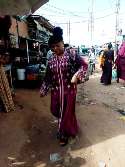 A medium shot captures a woman walking through a dusty market in Tempure, Nigeria. She is wearing a long, deep purple, embroidered kaftan with white and gold patterns on the sleeves and down the front. She carries a black handbag on her left shoulder and is wearing black sandals. Her hair is styled in a bun. In the background, other market-goers are visible. To her left, a man wearing a tie-dye t-shirt stands near a stall displaying various items, including buckets and woven baskets. Further back, a man on a bicycle is seen amidst other stalls and structures, with a cell tower prominently positioned against the bright sky. The ground is a mixture of dirt and debris.