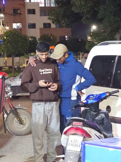 Two men stand beside a vehicle. One wears a brown hoodie and grey jeans, holding a phone. The other has on a blue jacket, grey pants, and a patterned hat. They appear to be interacting with each other near a scooter-like vehicle with a license plate visible. The background shows buildings, trees, and streetlights, suggesting an urban outdoor environment.