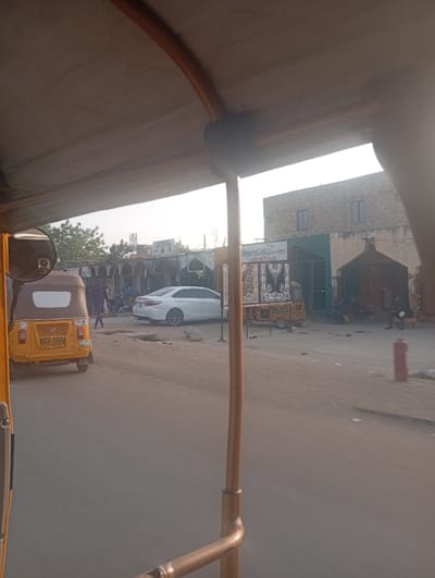 From the vantage point of a vehicle, potentially a rickshaw, the scene reveals a sunlit street. Another yellow three-wheeled vehicle is visible in the foreground. Further down the street, a white car stands out near storefronts. A building with a sign reading "Ayalaok.com LOGISTICS" is visible. The street appears to be sandy or dusty. Several other figures and vehicles are seen in the distance.