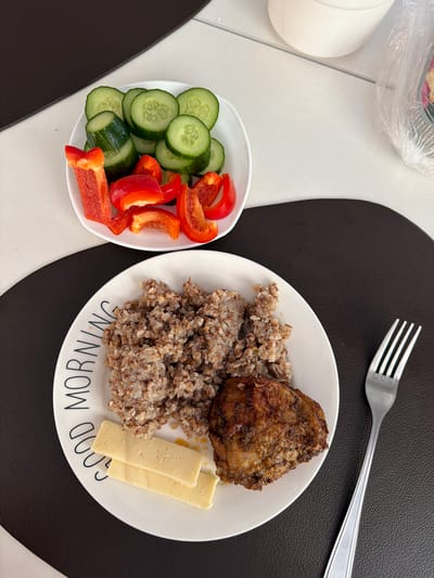 A close-up, overhead view shows a breakfast meal arranged on a table. In the foreground, a white plate with the words "GOOD MORNING" printed in a circular pattern around its edge contains a serving of buckwheat, a piece of cooked meat, and two slices of cheese. To the left of the plate, a white dish holds sliced cucumbers and chopped red bell peppers. A silver fork rests to the right of the plate, and the table is covered by a dark brown placemat. The background shows a white tiled surface and a partial view of a white bowl and a plastic-wrapped object. The scene is set in an indoor environment, likely a kitchen or dining area. There are no people visible in the image.