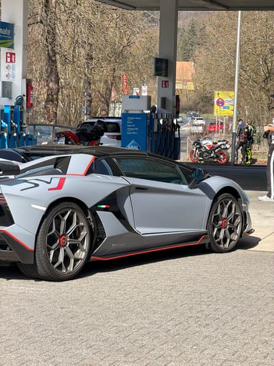 The image depicts a matte grey Lamborghini Aventador SVJ Roadster parked on asphalt pavement at an Aral gas station in Adenau, Germany, under bright daylight. The vehicle features red accents along its side skirts, rear diffuser, and a "SVJ" graphic on the rear quarter panel, along with an Italian flag emblem on the side air intake and multi-spoke wheels with red center locks.

In the mid-ground, a white SUV and a black motorcycle are parked behind the Lamborghini near blue Aral fuel pumps. Beyond the pumps, a road curves to the right, with several vehicles, including a red car, visible. A silver and red sportbike is parked on its kickstand beside the road.

Two individuals stand near the sportbike: one wears a black and yellow/lime green leather motorcycle racing suit, and the other wears a black and white leather racing suit. To the far right, a person with partially visible black and white tracksuit-style pants and white sneakers holds a dark object, possibly a camera or phone, toward the scene.

The background consists of bare deciduous trees on a hillside, suggesting late winter or early spring conditions. A light-colored building with an orange roof is visible amongst the trees. Yellow and blue road signs are present near the road curve. The gas station canopy structure, supported by pillars, casts shadows across the scene.