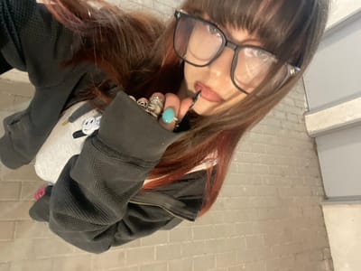 A female individual is depicted from an overhead perspective, looking directly into the camera, in the city of Raanana, Israel. She has long, brown hair with red highlights and bangs, and wears black-framed eyeglasses. Her right hand is raised, with multiple rings on the fingers, including a prominent turquoise-colored ring. The index finger's long, dark-painted nail is positioned near her lip. She is dressed in a dark grey waffle-knit hooded sweatshirt, partially revealing a white garment underneath that features a black and white smiley face graphic. A pink shoe is partially visible at the lower left. The environment consists of light-colored brick-patterned flooring and a light grey brick wall with a visible concrete column.