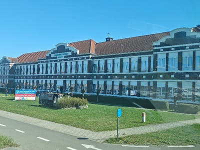 The image depicts the Sint-Martinusschool Zonneberg, a multi-story educational building in Genk, Belgium, observed from a public thoroughfare. The building is constructed of dark brick with light-colored trim around numerous evenly spaced rectangular windows, some featuring white interior blinds. It has a distinctive red-tiled hip roof with several gables and chimneys. A large sign above the main entrance clearly reads "ST. MARTINUSSESCHOOL ZONNEBERG".
In the foreground, a wide expanse of green grass separates the building from a paved road and sidewalk. A blue and white banner is positioned on the grass to the left of the building's center, displaying colorful text and graphics, including "inschrijven kan vanaf nu!" (registration is possible from now on!). To the right of the banner, a dark, weathered, tank-like metal sculpture is situated within a circular, low-hedged planter. Several bare-branched trees are scattered across the lawn in front of the building.
To the right foreground, a paved sidewalk runs parallel to the road. A blue circular sign with a white bicycle symbol, indicating a bicycle route, is visible on a green pole. A red and white marker post stands behind it. The road in the immediate foreground features white lane markings, including a solid line and a white arrow indicating forward movement. The sky is clear and bright blue. Two small, faint white contrails are visible in the upper right quadrant of the sky. A few individuals are faintly discernible near the left side of the building entrance.
