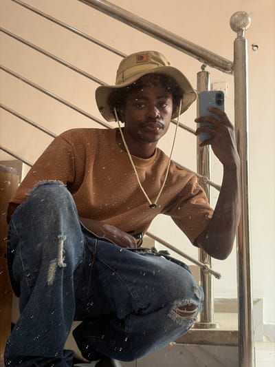 The image captures a young Black male crouching indoors in Igboefon, Nigeria, reflected in what appears to be a mirror covered with fine white specks, possibly dust or paint. He is actively taking a selfie using a light blue smartphone held in his raised left hand. The individual wears a tan boonie hat with a chin strap, a muted orange crew-neck short-sleeved t-shirt, and distressed blue denim jeans with large rips at both knees, all of which are visibly splattered with small white paint marks. He has a short afro hairstyle and a faint mustache, maintaining a neutral expression directed towards the camera. The background, visible through the reflection, consists of a light-colored wall and a multi-barred metallic staircase railing.