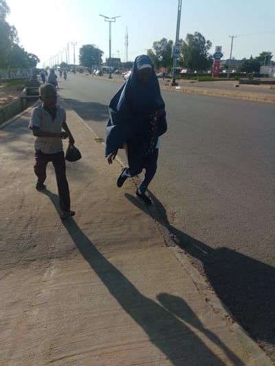 The image captures a street scene in Tumfure, Nigeria, likely in the late afternoon or early morning due to the long shadows. In the foreground, a young boy wearing a checkered short-sleeved shirt and dark trousers walks on a concrete sidewalk, carrying a black plastic bag in his right hand. His left hand is outstretched. To his right, a person in a dark blue abaya, which covers them from head to toe, is walking on the asphalt road, also casting a long shadow. The abaya appears to have a pattern on it, possibly embroidered. The road is paved with asphalt and has a curb separating it from the sidewalk. In the background, there is traffic, including several motorcycles and cars, and streetlights are visible along the road. Trees line the side of the road, and beyond them, buildings and communication towers can be seen. The sky is bright and clear.