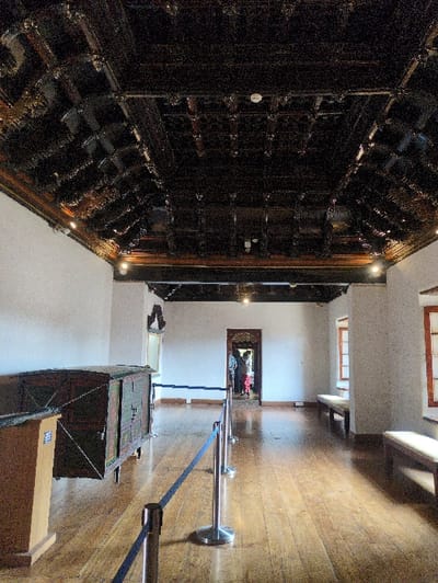 The image depicts a large, well-lit exhibition room within the Mattancherry Palace in Kochi, Kerala, India. The most striking architectural feature is the dark, intricately carved wooden coffered ceiling, adorned with numerous patterns and integrated spotlights. The walls are uniformly white, and the floor consists of polished light-brown wooden planks.
On the left side of the room, an antique dark wooden chest or cabinet, featuring distinct green and reddish decorative panels, is displayed. This artifact is positioned behind a visitor barrier composed of polished silver stanchions connected by blue velvet ropes, which extends into the mid-ground. Along the right wall, two plain wooden benches with light-colored cushions are situated beneath rectangular windows framed in dark wood.
At the far end of the room, a wide doorway with an ornate, dark carved wooden frame provides access to an adjacent area. Through this doorway, several indistinct figures, identified as visitors, are visible, some wearing brightly colored clothing, suggesting movement into or out of the room. The overall scene is static, captured from a slightly low-angle perspective, emphasizing the ceiling and the depth of the room.