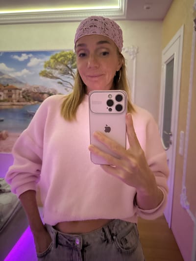 The image shows a woman in a pink knitted headband and a pink oversized sweater, holding a smartphone to take a selfie. She is wearing light-wash jeans and has one hand in her pocket. The background is a room with a wall mural of a landscape, a framed mirror, and a closed door. The lighting in the room appears to be a mix of white and purple. The woman is looking at the camera with a slight smile. The specific location in the city of Sinyavino, Russia cannot be determined from the image alone.