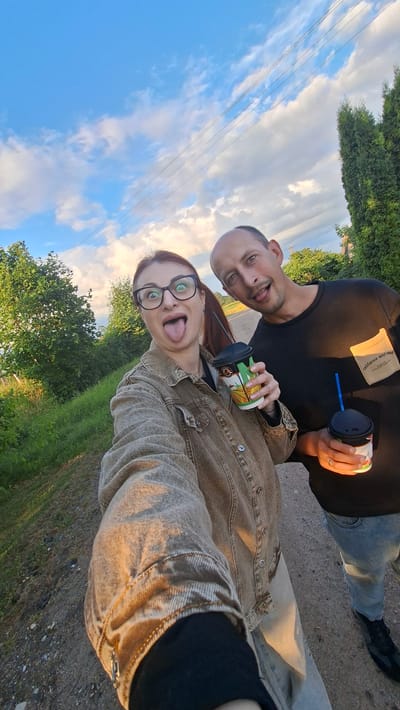 A selfie is taken by a couple outdoors on a dirt road. The woman in the foreground is wearing glasses and a denim jacket, sticking her tongue out at the camera. She holds a cup with a blue straw. Behind her, a man in a black t-shirt and blue jeans is also sticking his tongue out and holding a cup with a blue straw. Trees and greenery line the road, with a cloudy sky above. Power lines are visible in the distance. The setting appears to be a rural area within the Забалоцкі сельскі Савет, Belarus.