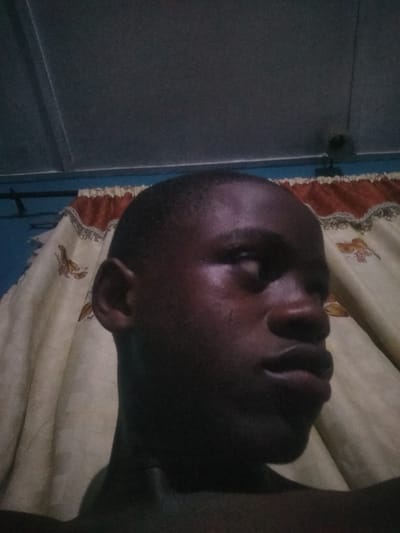 The image depicts a person with dark skin looking towards the right side of the frame. The person has short hair and is facing sideways. The background includes a tan-colored curtain with a red and white patterned border along the top, positioned behind the person. Above the curtain, there is a blue wall and a grey ceiling. A small black object is mounted on the wall near the curtain, near to the center of the image. There is insufficient evidence to determine the location is Epe, Nigeria.