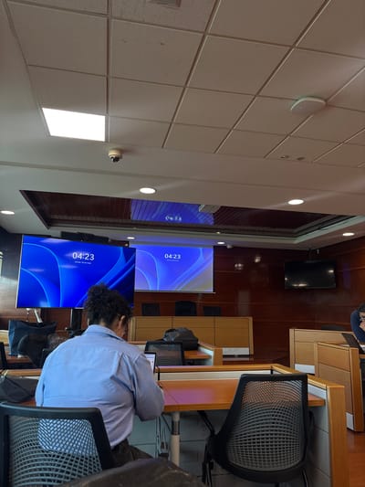 The image shows an indoor office space, likely a conference room or training center, in the city of Puebla, Mexico. Two large screens display the time "04:23" and Spanish text "Viernes, 05 de mayo", indicating it is Friday, May 5th. An individual with dark curly hair, wearing a light blue collared shirt, is seated at a desk, seemingly working on a laptop or tablet. The room has wood-paneled walls and a tiled ceiling with recessed lighting. A black flat-screen TV is mounted on the wall. The desks are made of light-colored wood, and chairs are black with mesh backs. The scene appears to be a professional or educational environment.