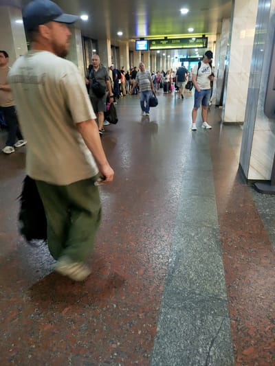 This image depicts the interior of a building that appears to be a transportation hub, possibly a train station or airport terminal, in Kyiv, Ukraine. The flooring is a speckled red and gray granite. A man in the foreground, wearing a light-colored t-shirt, green pants, and a blue baseball cap, is walking away from the camera, carrying a dark bag. He appears to be right-handed, as his right hand is visible and holding a small item. In the background, other people are walking in various directions, some carrying luggage. Overhead, there are fluorescent lights and a digital display board showing times and destinations, consistent with a transportation terminal. The overall atmosphere suggests a busy, public space.