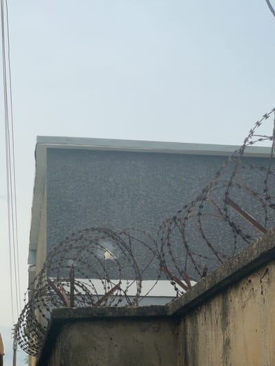 A concrete wall topped with barbed wire fences a building with a gray, textured exterior. The sky above is overcast. Several thin wires, possibly electrical or telephone lines, run vertically along the left side of the image. The scene appears to be an outdoor environment. The location is in the city of Narayi, Nigeria.