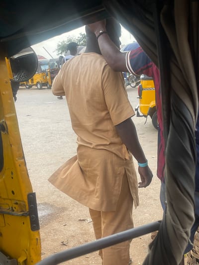 The image, captured within the bustling environment of Galadimawa Village, Nigeria, depicts a street scene dominated by a yellow auto-rickshaw, locally known as a "Keke." The foreground is occupied by the interior of another Keke, framing the view. A man, seen from the back and side, is dressed in a tan, loose-fitting traditional outfit, with a bracelet on his left wrist. He appears to be interacting with someone to his right, whose arm, adorned with a striped sleeve, is visible. In the background, other Kekes and individuals are discernible, suggesting a typical day in this Nigerian locale. The ground is unpaved, indicative of a rural or semi-urban setting.
