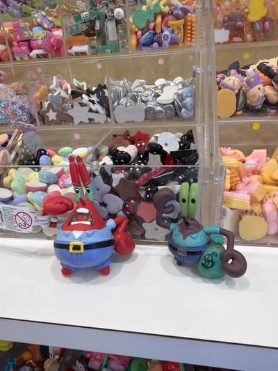 The image shows a close-up, eye-level view of a shelf display at a store, likely in Milan, Italy, based on the text on a label in the lower left corner. The primary focus is on two character figurines that appear to be from the Spongebob Squarepants cartoon: Mr. Krabs on the left and Squidward Tentacles on the right. Both figurines are in a standing, dynamic pose. Mr. Krabs is depicted with his mouth open, red arms extended, and wearing a blue uniform with a yellow belt. Squidward is shown with his green head and arms, holding a green money bag with a dollar sign on it.
Behind the figurines, a transparent plastic organizer is filled with a variety of colorful craft supplies and decorative items. These include small, brightly colored plastic shapes such as stars, hearts, and other abstract designs, as well as what appear to be miniature food items like donuts and candies. Some of the compartments in the organizer also contain miniature bottles and other small trinkets. The lighting is bright and evenly distributed, suggesting an indoor retail environment. The overall impression is of a display designed to attract customers looking for craft materials or novelty items.