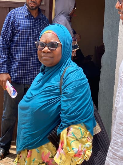 A medium shot of a woman wearing a blue hijab and sunglasses, standing next to a man in a blue checkered shirt, holding a rolled-up stack of papers. A yellow floral print bag is seen behind her. The location is identified as Sabon Tasha, Nigeria.