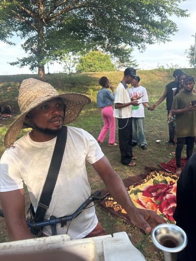 A group of people are gathered in an outdoor setting in Ihiagwa, Nigeria. The foreground features a man wearing a straw hat and a white t-shirt, with a strap across his chest. He is holding a metallic object with a round opening towards the viewer. Behind him, there is a group of people standing on the grass. Some are holding phones and appearing to take pictures. There is also a patterned blanket on the ground. The environment includes green grass, trees, and a slightly overcast sky.