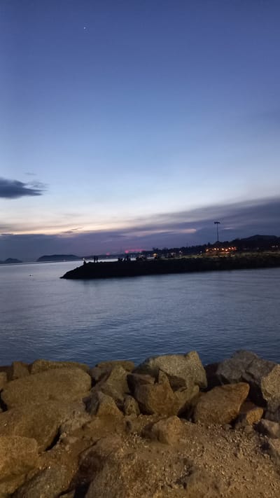 The image shows a twilight scene at the sea. In the foreground, there is a pile of large, rough rocks and gravel. Beyond the rocks, a calm body of water stretches out to the horizon. On the water, in the middle ground, a dark, rocky outcrop or jetty juts out into the sea. Silhouettes of several people can be seen standing on this outcrop. In the distance, the shoreline is visible with hills and buildings, illuminated by lights. A single street lamp with two bulbs stands prominently on the jetty. The sky above is a gradient of deep blue at the top, fading to lighter blues and purples near the horizon, with scattered clouds. There are two bright points of light in the upper sky, likely stars or planets. The overall atmosphere is peaceful and serene, capturing the transition from day to night. The location is specified as being in the Ban Tai Subdistrict, Thailand.