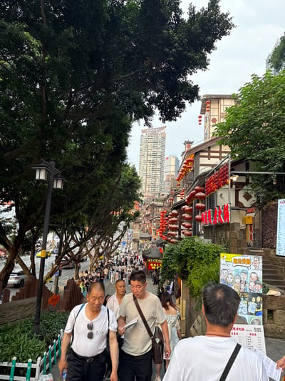 The image depicts a bustling street scene in Yuzhong District, Chongqing, China. The architecture consists of traditional-style buildings adorned with rows of red lanterns, mixed with a modern skyscraper in the background. A crowd of people fills the street, some pausing to read menus or look at advertisements. Several individuals are prominently visible in the foreground, appearing to be tourists or locals, dressed in casual clothing. Trees and street lamps line the street, adding to the urban landscape.