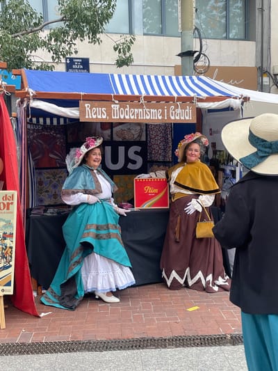 The image shows two women dressed in vintage or historical costumes standing behind a stall at a market or fair. A sign reads "Reus, Modernisme i Gaudí," suggesting a theme of modernism and the architect Antoni Gaudí. The stall is decorated with blue and white striped fabric. Another sign on a nearby building reads "CARRER DE LA GOLETA." A red box labeled "Plim" is visible on the stall, as is a poster with the words "del or." Based on the street name and the reference to Reus, this likely takes place in or has connections to Reus, Spain.