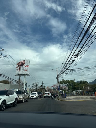 The image was taken from the driver's seat of a car, looking forward. The road is paved and has a line of traffic in front. The image was captured in San Rafael, Costa Rica, located at latitude: 9.938, longitude: -84.153. The sky is blue with white fluffy clouds. In the foreground, the car is stopped in traffic, and there is a line of cars in front. There are trees and power lines on both sides of the road. In the background, a large billboard is visible on the left side of the road. The billboard is displaying a large advertisement for the clothing company H&M.  The road is relatively empty except for the cars in traffic, and the traffic flow is moving slowly.  There are numerous power lines in the air, and the scenery is urban, but a few trees indicate nearby green spaces.