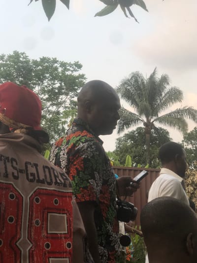 The image depicts an outdoor scene in Awka Etiti, Nigeria, featuring multiple individuals under an overcast sky. In the foreground, the back of a person wearing a beige top with red and white lettering "TS-GLOR" and "88," and a red baseball cap with a dark fabric band, is visible on the left. To their right, a bald, dark-skinned male in a black, orange, green, and white floral-patterned short-sleeved shirt is shown in profile, holding a black smartphone in his right hand and looking at its screen. A dark camera or recording device is strapped to his left side. Further to the right, a partially obscured dark-skinned male in a light-colored collared shirt is visible in profile. The top of another dark-skinned, closely shaven head appears in the bottom right foreground. The background features lush tropical vegetation, including large trees and a prominent palm tree, beneath a light gray, cloudy sky. A light brown corrugated metal fence or wall is present behind the mid-ground figures, with various green plants and some out-of-focus light-colored objects in front of it. Overhanging green leaves are visible in the top portion of the frame.
