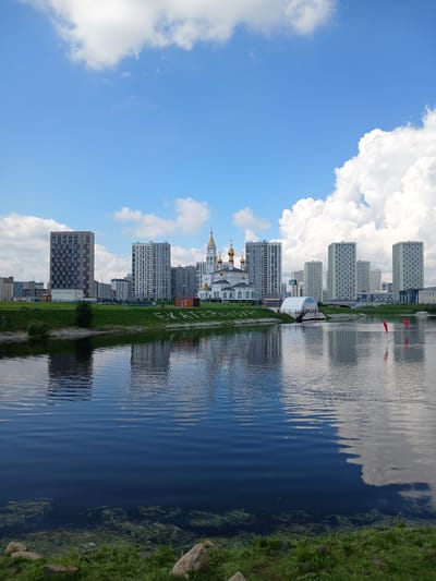 The image, taken in Yekaterinburg, Russia, depicts a cityscape reflected in a body of water. The scene is oriented vertically, suggesting the photo was taken sideways and then rotated.
Dominating the left side of the frame is a bright blue sky filled with fluffy white clouds. These clouds appear to be illuminated by the sun, indicating a daytime setting, likely mid-morning or afternoon given the brightness.
The right side of the image features a waterfront with a grassy bank on the far right. A dark body of water, likely a river or lake, occupies the central portion, mirroring the sky and the buildings. The water is slightly rippled, creating a distorted reflection of the surrounding elements. Submerged aquatic plants are visible in the clearer sections of the water near the bank.
The cityscape consists of numerous tall, modern buildings, predominantly residential high-rises, constructed with light-colored facades. In the foreground, a white building with a distinctive rounded, glass-like dome is visible. Further to the left, a building with golden domes and crosses suggests the presence of a church or cathedral, a common feature in Russian cities. A pathway runs alongside the water, separating the buildings from the grassy embankment. Two red kite-like objects are visible in the water, suggesting some recreational activity.
There are no visible people in the image. The overall atmosphere is serene and tranquil, showcasing a harmonious blend of urban architecture and natural elements under a clear, cloudy sky.