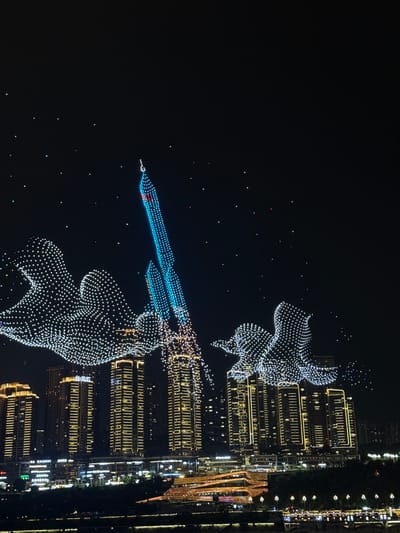 This is a night-time aerial photograph of a drone light show over a city skyline. The subjects are numerous drones, illuminated with white and blue lights, forming abstract shapes and figures in the sky. Two prominent figures, resembling abstract humanoids or mythical creatures, are visible, one at the top right and another at the bottom. Between them, a long, blue, beam-like formation of drones is depicted, possibly representing a projectile or a pathway. The drones are creating intricate patterns against the dark night sky, with scattered colorful lights adding to the display.
In the background, the city lights of Yuzhong District, China, are clearly visible. Tall buildings with illuminated windows create a dense urban landscape, stretching vertically along the right side of the image. The lights from the city, including streetlights and building illumination, reflect on what appears to be a body of water, suggesting the show is taking place near a waterfront.
The activity taking place is a drone light show, an organized event likely for entertainment or celebration. The time of day is clearly night, given the dark sky and the prominence of artificial lights. No specific weather conditions are discernible, but the clarity of the drone formations and city lights suggests clear weather. The overall mood conveyed is one of awe and wonder, characteristic of spectacular light displays. There is no visible text in the image.
