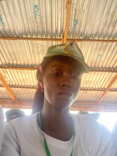 A medium shot selfie captures a young woman wearing a white t-shirt and a green cap. The cap features a circular emblem with a depiction of a person in profile, and it is adorned with white and green stripes. She has braided hair with red accents and wears red earrings. The background reveals a corrugated metal roof supported by wooden beams. The overall scene suggests an outdoor, possibly informal setting, consistent with the architecture observed. The image is shot from a low angle, looking up towards the subject and the roof. The lighting is bright, suggesting it is daytime, and there is a lens flare in the upper right portion of the image. The woman's expression is neutral. The context provided places this scene in the city of Jimeta, Nigeria.