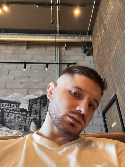 The image depicts a male individual, appearing to be in his 20s or 30s, captured in a head-and-shoulders frame. He has dark, short hair with faded sides and a longer top, styled back. A short, trimmed beard and mustache are visible on his face. He is wearing a light-colored, possibly cream or pale yellow, crew-neck t-shirt. His left ear features an earring. The individual is looking directly at the camera with a neutral expression.
The scene is indoors, located in what appears to be a commercial establishment, specifically in Bretton, United Kingdom, as stated. The background features an industrial-style interior. The ceiling is dark, with exposed silver ventilation ductwork running horizontally and exposed silver conduits running vertically and horizontally. Track lighting with multiple black spotlights and two brighter circular lights illuminate the space. A small green indicator light is visible on the ceiling.
The left wall has a light grey brick pattern, overlaid with a black and white mural depicting architectural structures, resembling an old city gate or historical buildings. The right wall consists of dark, vertically corrugated paneling. A black-framed door with a reflective or frosted panel displays the word "TOILET" in white lettering on the right side of the frame. A black security camera or speaker is mounted on the brick wall segment to the right of the main ventilation duct. The individual appears to be seated on a brown upholstered surface, partially visible at the bottom right. No other individuals or interactions are present.