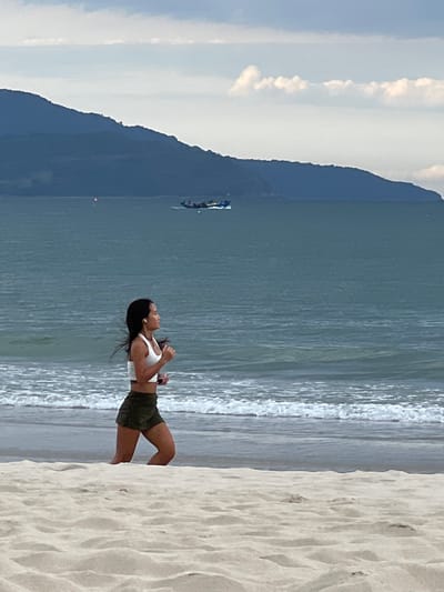 The image shows a woman standing on a sandy beach, facing the ocean. She is wearing a white crop top and olive green shorts. Her hair is dark and appears to be blowing in the wind. She is looking up and to her left, with her right arm raised. There are waves gently breaking on the shore in front of her. In the distance, across the water, a large, dark mountain looms, with clouds scattered across the sky above it. There is also a small boat visible on the water in front of the mountain. The sand in the foreground is light-colored and appears to have footprints on it. The lighting suggests it is daytime, likely late afternoon or early morning due to the soft light and the colors of the sky. The scene evokes a sense of peace and contemplation.