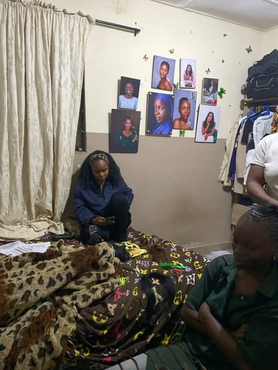 In a room in Durumi, Nigeria, a young woman with dark, curly hair sits on a bed covered with a leopard-print blanket and a brown blanket with a Louis Vuitton pattern. She is wearing a blue jacket and black pants and is looking down at her phone. On the wall behind her are several framed portraits of women. To her right, another young woman with braided hair, wearing a green shirt, is also sitting on the bed, her arms crossed. In the background, clothes hang on a rack, and luggage is stacked on a shelf. The room appears to be a bedroom.