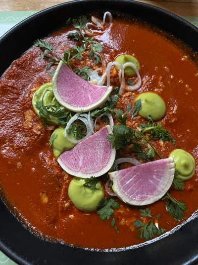 A close-up, overhead shot displays a plate of Mexican cuisine in San Bernardino Tlaxcalancingo, Mexico. The dish features a vibrant red sauce covering the base, which appears to contain enchiladas or a similar type of filled tortilla. Toppings include dollops of light green avocado cream, slices of pink-purple watermelon radish, thin slivers of white onion, and sprigs of fresh cilantro. The dish is served in a deep, black, possibly cast-iron pan.