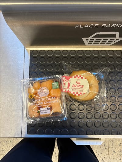 The image captures a close-up shot of a conveyor belt at a supermarket checkout. On the belt are two packaged food items: a salmon bagel from Marks & Spencer (M&S) and a container of cantaloupe melon chunks, also from M&S. A sign indicating "PLACE BASKET" is visible on the side of the conveyor belt. The black surface of the conveyor belt has a patterned texture with circular indentations. A person wearing black trousers is partially visible to the right of the frame. Based on the M&S branding and the overall setting, the location is likely a supermarket in London, United Kingdom.