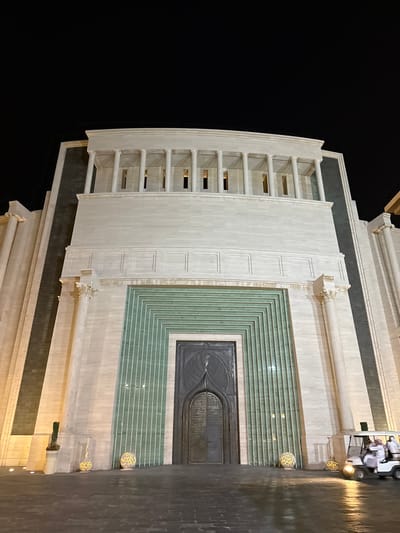 The image shows a building facade at night. The building is located in Doha, Qatar. The facade is predominantly white, with a large decorative door framed by geometric patterns in a greenish-blue hue. A series of open, stacked horizontal architectural elements are visible on the left side of the facade. To the right, a golf cart with several people inside sits along what seems to be a paved walkway. Decorative, spherical light fixtures are placed along the walkway. The background is predominantly dark, suggesting it is night.