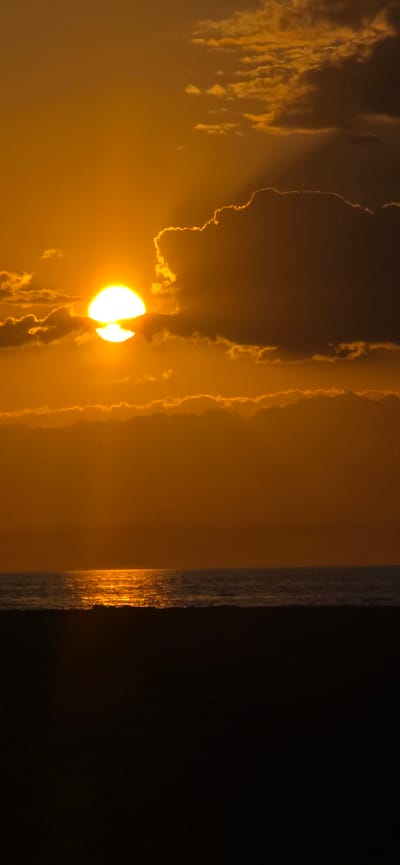 The image depicts a sunset over the sea. The sun, a bright orb of light, is partially obscured by dark clouds. The clouds are illuminated by the setting sun, creating a dramatic contrast between light and shadow. The sea is calm, with a shimmering reflection of the sun on its surface. The foreground is a dark silhouette, likely of a beach or land. The scene is set in Boz Beshik, Kyrgyzstan.