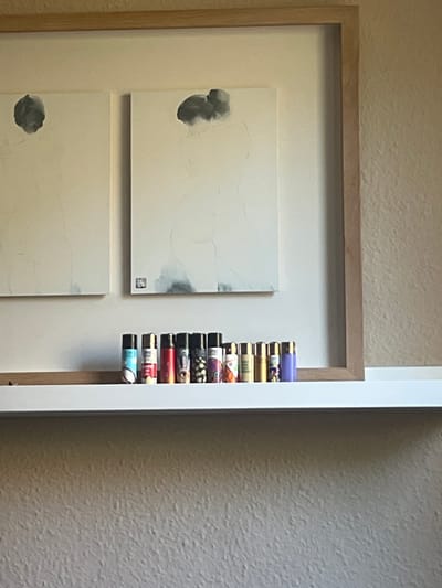 This media file shows a close-up view of a framed artwork with a stack of objects beside it. The artwork appears to be abstract, with two white panels featuring light grey ink washes. The top panel has a splotchy grey form on the left, and the bottom panel has a similar grey form on the left. A small, dark square is visible in the lower right corner of the top panel. The artwork is framed with a light-colored wood.

To the right of the artwork, a vertical stack of colorful cylindrical objects is visible. These objects appear to be lighters, with various patterns and colors, including purple, yellow, green, red, and blue. They are neatly arranged in a row.

The background consists of a textured wall with a light beige or cream color. The lighting suggests it is daytime, with soft illumination casting subtle shadows. The setting appears to be indoors, possibly a home or gallery space, given the framed art and the way the objects are displayed. There is no visible text in the image. The overall impression is one of organized display, with the art providing a focal point and the stack of lighters adding a contrasting element of color and texture. The location context of Madrid, Spain, does not provide any specific visual cues within this cropped image.