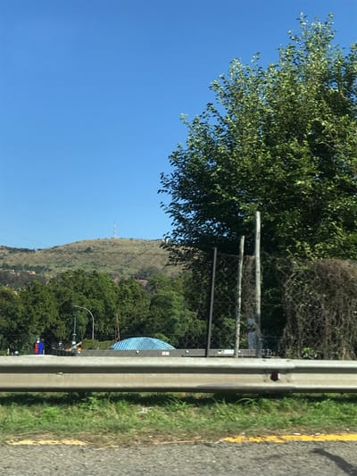 A roadside view with a highway barrier dominating the foreground. Lush green foliage cascades from the top left, meeting a chain-link fence. Beyond, a grassy hill rises under a clear blue sky. Trees dot the landscape, obscuring structures at the hill's base. A communication tower stands atop the hill. The road's edge, defined by a yellow line and green grass, contrasts with the gray asphalt.