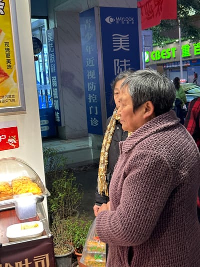 This image captures an urban street scene in Jiangjin, China, during the evening, characterized by artificial illumination. In the foreground, an older woman with short, graying hair and a dark purple knitted cardigan is positioned with her right side visible, looking towards the left. Her right hand holds a clear plastic bag containing various items. Behind her and slightly to the left stands another older woman, partially obscured, with dark hair, a dark jacket, and a distinctive patterned yellow, white, and brown scarf, also appearing to look in the same direction.

To the immediate left of the foreground woman is a mobile food display unit. It features a clear domed cover over multiple golden-brown baked goods, likely pastries. Below this, a small white rectangular dish contains a single yellow, elongated snack item, and an adjacent container holds white, granular material. A partial Chinese sign is visible beneath the display.

In the mid-background, two prominent blue vertical signs are visible. The rightmost sign displays "MAYLOOK" with an eye logo and "美陆眼科" (Maylook Ophthalmology), followed by "青少年近视防控门诊" (Youth Myopia Prevention and Control Clinic). The leftmost blue sign, partially obscured by a poster, also reads "美陆眼科" and "弱视训练" (Amblyopia Training). Between the food stand and the clinic entrance, small potted green plants are arranged on the ground.

Further in the background, a blurred urban environment includes a green neon sign reading "CBEST," other indistinct businesses, and the silhouettes of additional pedestrians and vehicles, suggesting an active commercial thoroughfare. A red flag is partially visible in the upper right corner of the frame.