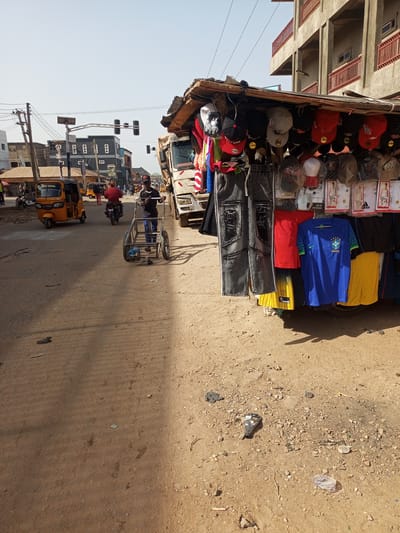 The image depicts a street scene in Maiburiji, Nigeria. The road is unpaved and dusty. A roadside stall displays clothing items, including hats, jeans, shorts, and jerseys. A man is pulling an empty metal cart down the street. There is a tricycle and a motorcyclist behind him. A white truck is parked near the stall. Traffic lights are visible in the distance. Buildings are on the other side of the street. The sky is clear.