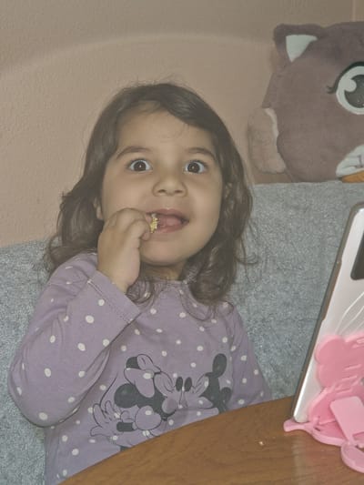 A young girl with brown hair and eyes is lying on a gray blanket. She is wearing a lavender long-sleeved shirt with white polka dots and a Mickey Mouse design. The girl is holding a small piece of food, possibly a puffed grain, in her hand and putting it in her mouth. To the left of the girl is a plush, cartoon-like animal toy with large, wide eyes. Above the girl is a pink object resting on a white surface, possibly a phone or tablet. The background consists of a pink wall and part of a wooden table. The specific location in Byala Slatina, Bulgaria, is not discernible from the image.