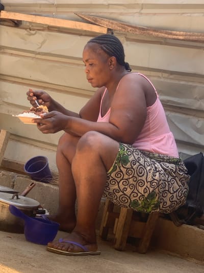 A woman with braided hair and a pink top crouches, eating cake with a fork. She wears a patterned skirt and flip-flops. Nearby are a blue cup and a blue scoop. A cooking pot with a lid and a metal stand are also visible. The background consists of corrugated metal and stacked wooden planks.